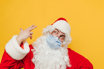 Santa Claus in a protective medical mask shoots himself in the head with a finger on a yellow background. Christmas in the coronavirus pandemic.