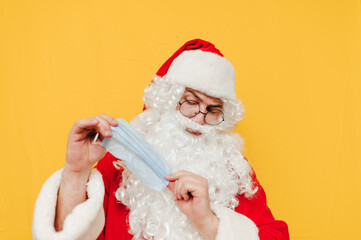 Obraz na płótnie Canvas Funny Santa Claus isolated on a yellow background with a protective mask against coronavirus in his hands. Santa Claus in a coronavirus pandemic uses a medical mask.