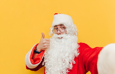 Positive Santa Claus takes a selfie on a yellow background and shows a thumbs up, looks at the camera and smiles. Christmas selfie from Santa