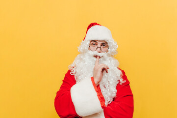 Fototapeta na wymiar Serious Santa Claus shows a sign of silence and looks intently at the camera on a yellow background. Santa shows a finger to the lips gesture Shhh