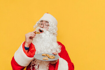 Santa Claus eats chocolate Christmas cookies on a yellow background with a plate. Christmas and New Year concept.