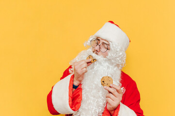 Fototapeta na wymiar Portrait of happy santa claus eating sweet chocolate chip cookies with closed eyes on yellow background. Santa eating christmas sweets, isolated.