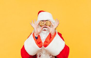 Funny santa with chocolate chip cookies in hands holding eyes near yellow background. Santa Claus and Christmas cookies, isolated. New Year