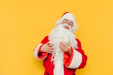 Funny Santa enjoys milk from a glass with his eyes closed on a yellow background. Funny man in...