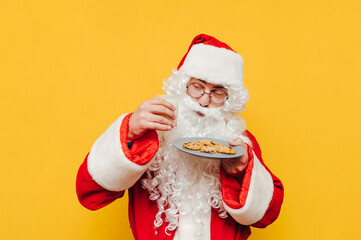 Funny Santa Claus stands on a yellow background with a plate of chocolate chip cookies and a glass of milk in his hands on a yellow background. Christmas and New Year concept