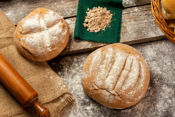 
Homemade round country bread with flour on wooden board with rolling pin, eggs, yeast cloth and rustic cloth with logs in the background