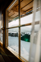 Wooden Cottage House Window. Beautiful snowy winter morning behind the chalet window. Beautiful mountain view. Sunshine and sparkling snow on fir trees in the cabin backyard.