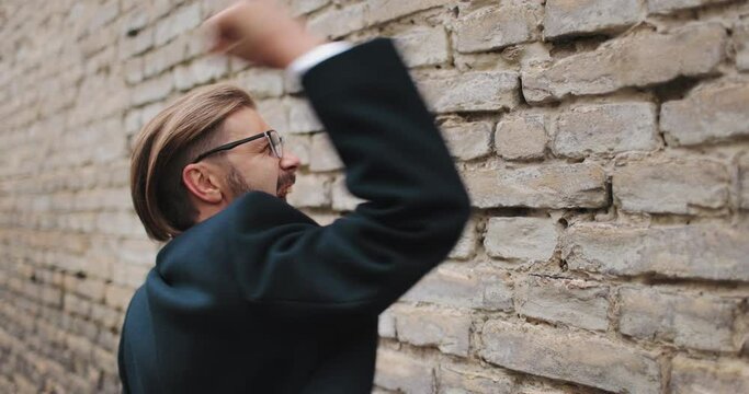 Angry bearded businessman pounding brick wall with fists and emotionally screaming outdoors. Man wearing eyeglasses and formal business clothing.