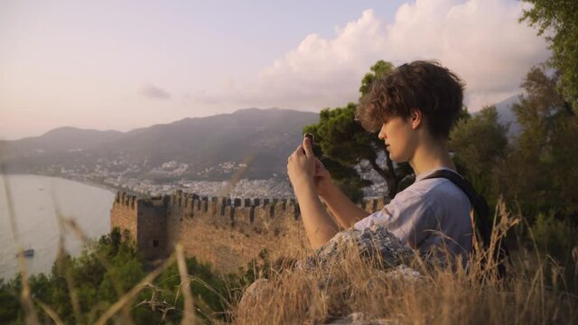 Medium shot of a young man using smartphone at sunset, fortress and mountains in the background, Alanya, Turkey