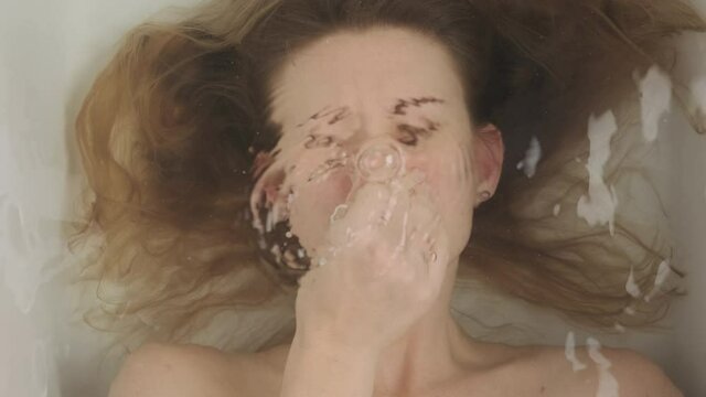 Woman lies in bathroom underwater, top view. lady holds her nose with her hand so that water does not get there, then emerges from under water in bath.