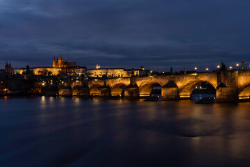
Charles Bridge on the Vltava river and statues on it and lit street lights and light from lamps at night in the center of Prague in the Czech Republic and there are clouds in the sky