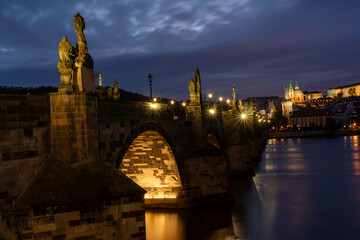 Fototapeta premium Charles Bridge on the Vltava river and statues on it and lit street lights and light from lamps at night in the center of Prague in the Czech Republic and there are clouds in the sky