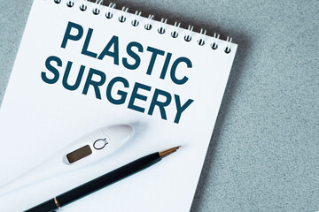 PLASTIC SURGERY is written in a white notebook on a gray table with a pen and a thermometer. Medical concept.