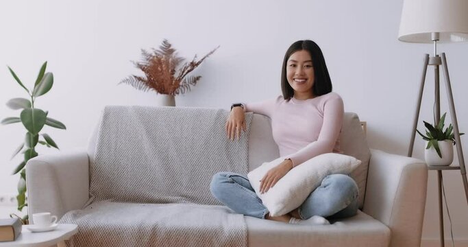 Domestic comfort. Happy asian woman sitting on sofa at home at laughing at camera