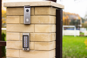 Silver intercom call panel with blue number buttons and a video camera on a brick beige fence post of a private house