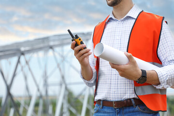 Professional engineer with plan and walkie talkie near high voltage tower construction outdoors, closeup. Installation of electrical substation