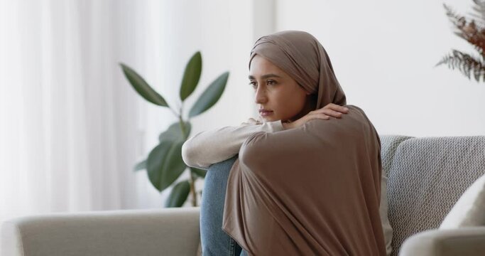 Depression concept. Upset muslim woman in hijab sitting alone on sofa at home, thinking about personal problems