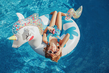 Cute adorable girl in sunglasses with drink lying on inflatable ring unicorn. Kid child enjoying having fun in swimming pool. Summer outdoors water activity for kids. View from above overhead. - 390264430