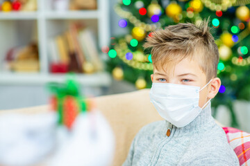 A boy with a medical mask on his face sits at home on the sofa against the background of a Christmas tree and looks at Santa's hands holding out a gift to him. Holidays during the coronavirus pandemic