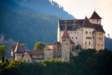 Fototapeta na wymiar Stone fortified Gutenberg Castle among green trees on top of hill on background with mountains and cloudy sky, Balzers, Liechtenstein