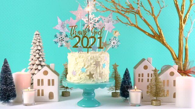 Happy New Year's Eve celebration cake on cake stand in blue white and gold theme decorated with stars and humorous, Thank God It's 2021, cake topper. Full party table setting with burning sparkler.