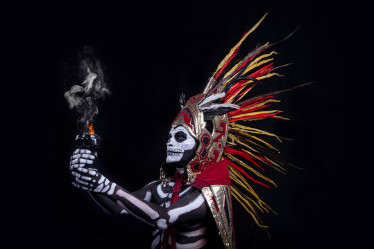 hombre latino , catrin de dios azteca, calavera prehispanica, con saumerio e incienso y fuego, portado penacho de plumas amarillas y rojas en fondo negro, dia de muertos, 1 de noviembre, calaca,