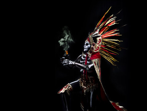 hombre latino , catrin de dios azteca, calavera prehispanica, con saumerio e incienso y fuego, portado penacho de plumas amarillas y rojas en fondo negro, dia de muertos, 1 de noviembre, calaca,