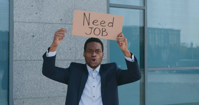 Desperate african american man in formal suit holding placard NEED JOB and shouting near office building, tracking shot