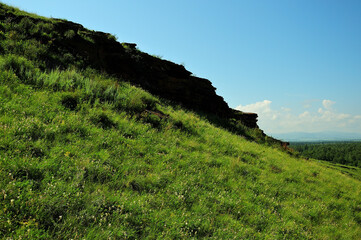 A flat rock mass at the top of a tall green hill under a clear summer sky.