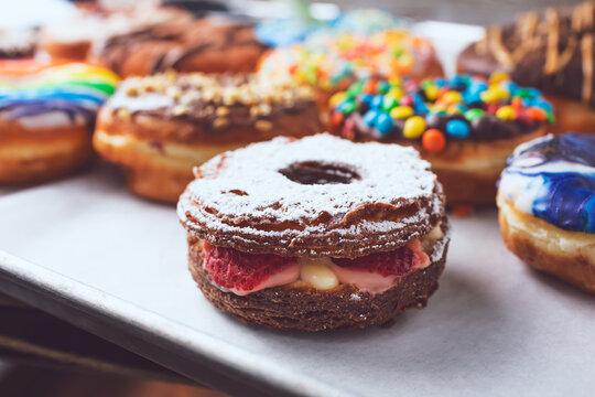 A view of a filled chocolate cronut among a baking sheet of assorted donuts.