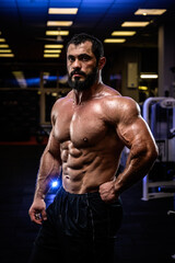 muscular strong young bearded caucasian male with powerful body in dark gym with blue and yellow lights