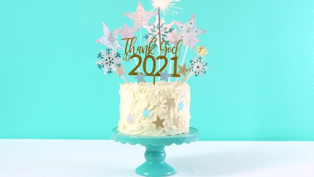 Happy New Year's Eve celebration cake on cake stand in blue white and gold theme decorated with stars and humorous, Thank God It's 2021, cake topper decoration with burning sparkler.