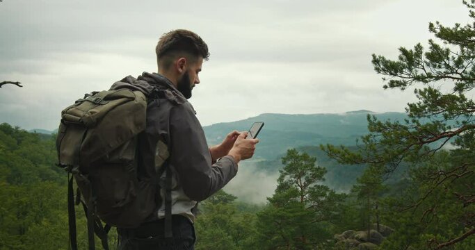 A man is standing on top of a mountain and typing a message on his smartphone. He has a large backpack on his shoulders. Hiking in the mountains. Top of the mountain. 4K DCI