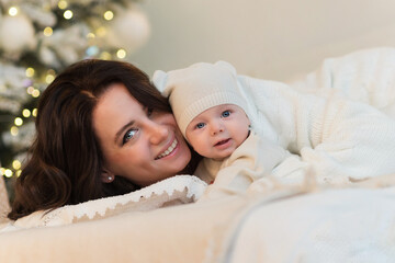 Mom and son are lying on bed in bright bedroom and are smiling happily. Woman hugs kid boy tightly. The concept of a Christmas morning, home comfort, scandy style. Merry Christmas and Happy New Year.