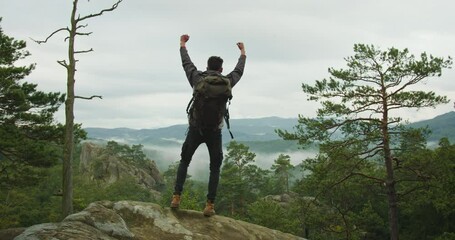 A man is going to the top of a cliff and looking at the landscape. He is raising his hands up and happy. He is carrying a large backpack. Hiking in the mountains. 4K DCI