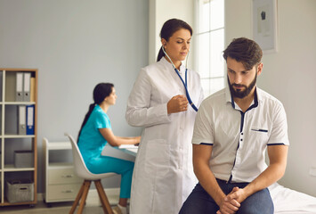 Female doctor in uniform listening to breath of young male patient through stethoscope