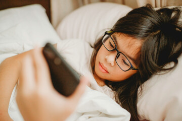 Asian child lying on bed playing smartphone.Chinese girl wearing glasses.Kid doing online learning in bedroom.Young female holding and reading through device.Tablet addiction. Bedsheet and pillows.