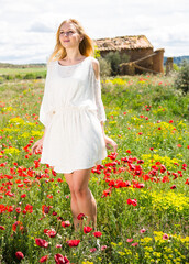 Naklejka na ściany i meble Beautiful young female in white dress walking through a poppy field of wild flowers