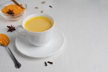Fotobehang Cup full of golden or turmeric milk which is healthy, healing drink having anti-inflammatory properties served on plate with spoon of curcuma on white wooden background. Image with copy space © Elena