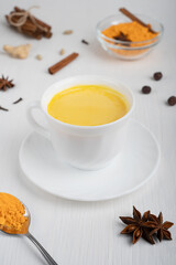Obraz na płótnie Canvas Cup full of golden or turmeric milk which is healthy, healing drink having anti-inflammatory properties served on plate with spoon of curcuma and anise on white wooden background. Vertical orientation
