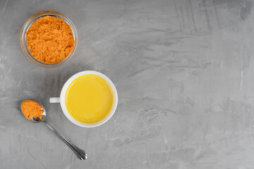 Top view of cup full of golden or turmeric milk which is healing drink having anti-inflammatory properties served with spoon of curcuma on gray concrete background. Image with copy space, horizontal