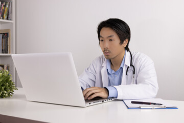 Young Asian Doctor Man in Lab Coat or Gown with Stethoscope Using Laptop Computer on Doctor Table in Office