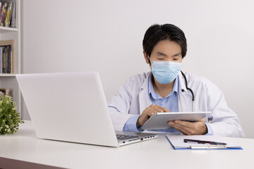 Young Asian Doctor Man in Lab Coat or Gown with Stethoscope Wear Face Mask Using Digital Tablet on Doctor Table in Office