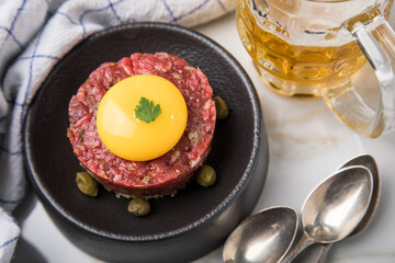 Gourmet Rindertatar Beef Tatar roh aus Filet fettarm, Kapern, Eigelb, Zwiebeln auf Toast Brot und schwarz Teller mit Bier, Löffel, Geschirrtuch und Marmor Hintergrund hell