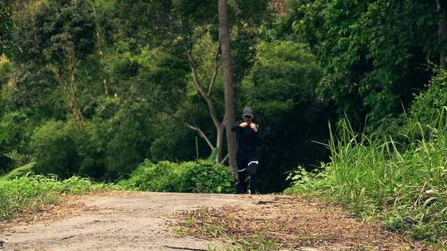 An athletic caucasian man in black workout clothes runs and punching in a forest on a sunny summer morning. Slow motion video.