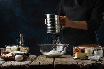 Fototapeta na wymiar The chef in black apron sifts flour into glass bowl for preparing dough. Backstage of cooking waffle on rustic wooden table with ingredients on dark blue background. Frozen motion. Sweet dessert.