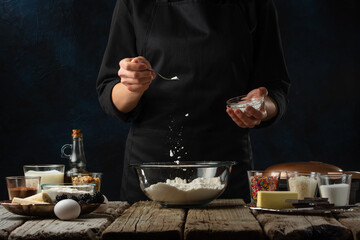 The chef in black apron pours salt into glass bowl for preparing dough. Backstage of cooking waffle on rustic wooden table with ingredients on dark blue background. Frozen motion. Traditional recipe.