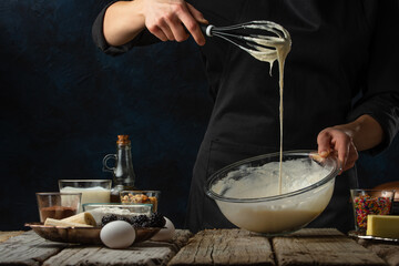 The chef in black apron kneads the dough for cooking waffle on rustic wooden table with ingredients on dark blue background. Frozen motion. Baked concept.