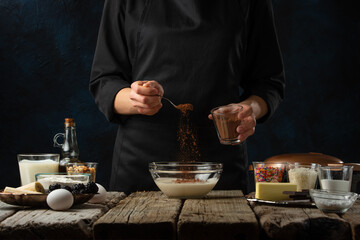 The chef in black apron pours cocoa into glass bowl for making dough. Backstage of cooking waffle on rustic wooden table with ingredients on dark blue background. Frozen motion. Traditional recipe.