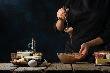 The chef in black apron testing the dough for cooking waffle on rustic wooden table with ingredients on dark blue background. Frozen motion. Bakery concept.